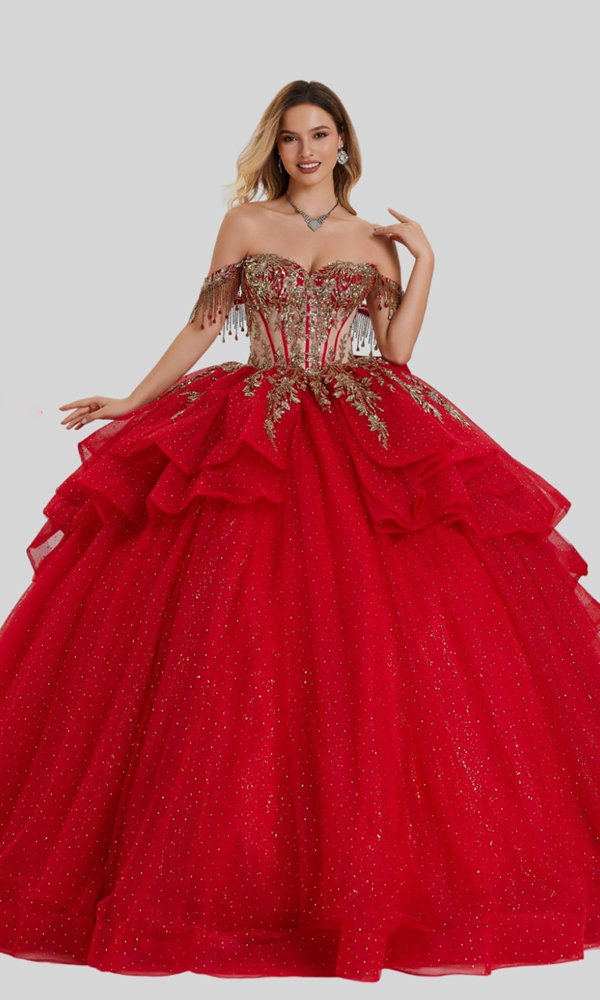 A woman stands against a plain grey background wearing the Quinceanera Dress 26417 by Stella Couture, a striking red off-the-shoulder ball gown featuring a gold-embellished corset bodice and a layered glitter tulle skirt.